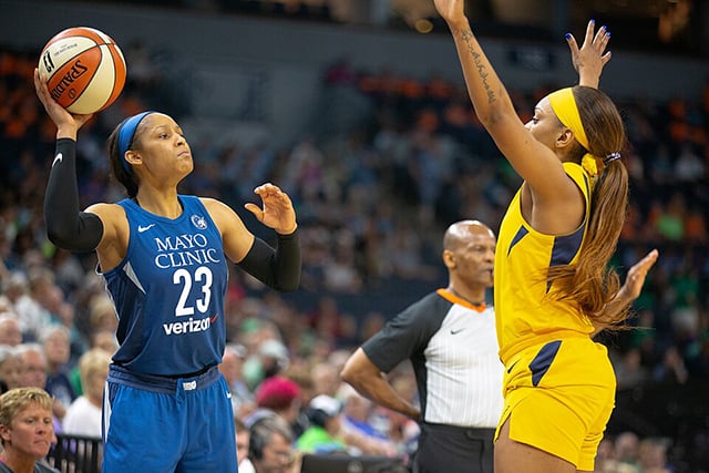With fans out of focus in the background, a basketball player in blue, holding a ball, faces off against a player in yellow, with a referee behind her.